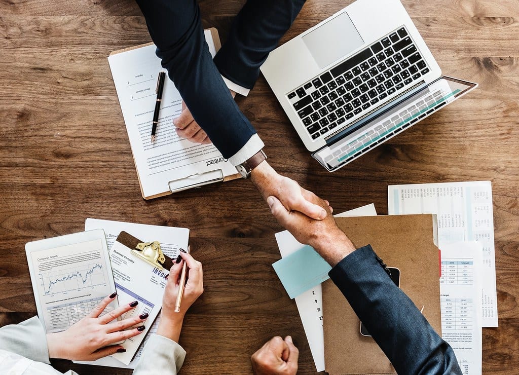 Two people shaking hands over a desk with paperwork and a laptop, signing a business agreement