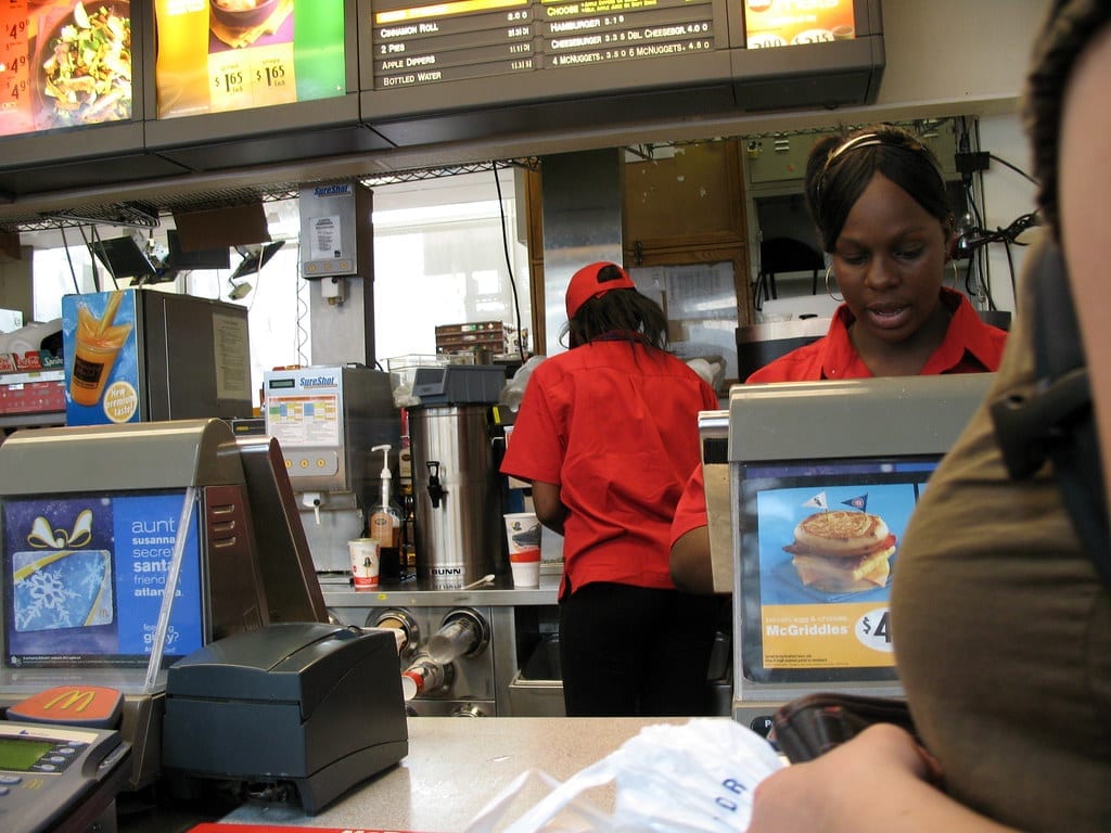 McDonald's crew members working the front counter and register, with the menu board and a McGriddles ad visible above