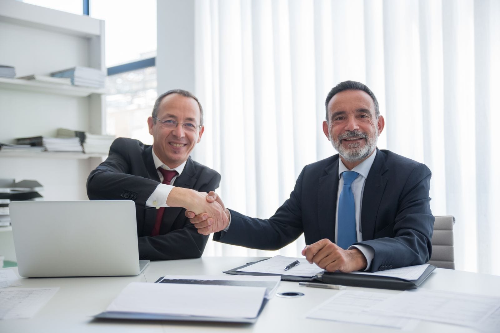 Two business partners shaking hands over a signed agreement and laptop at an office desk