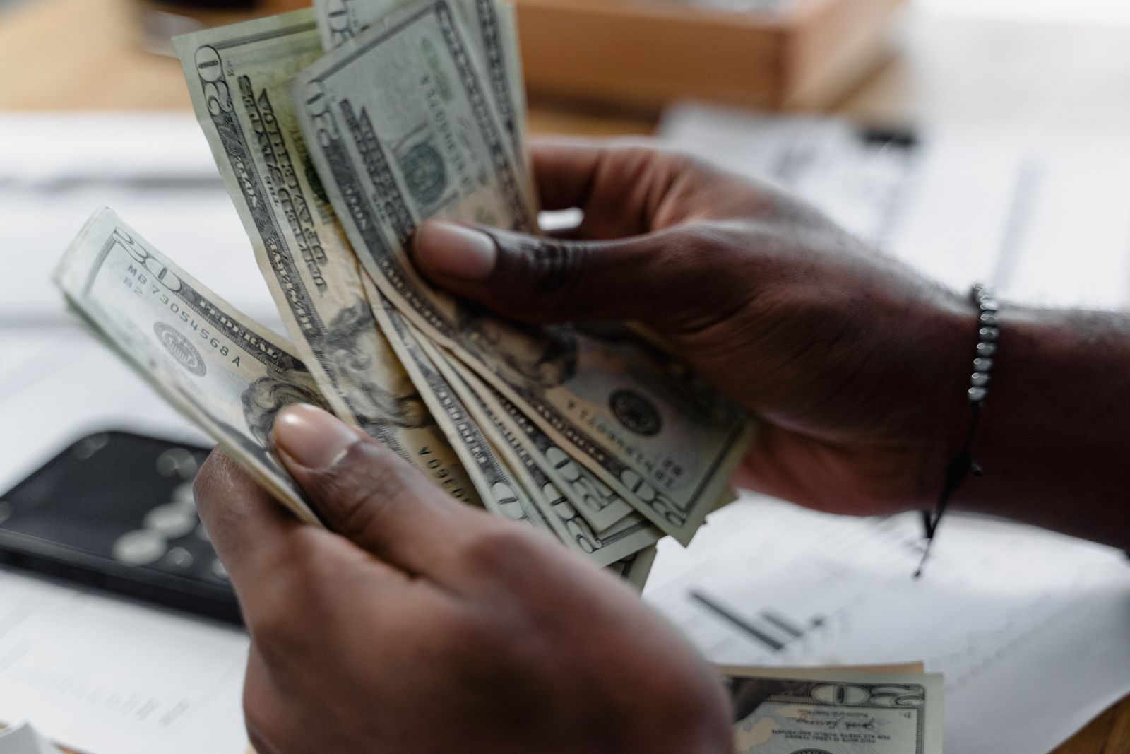 Hands counting a fan of twenty-dollar bills over financial paperwork on a desk