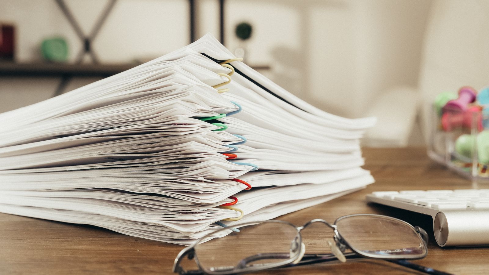 A tall stack of papers held with coloured clips on a wooden desk, a pair of reading glasses resting in front of it