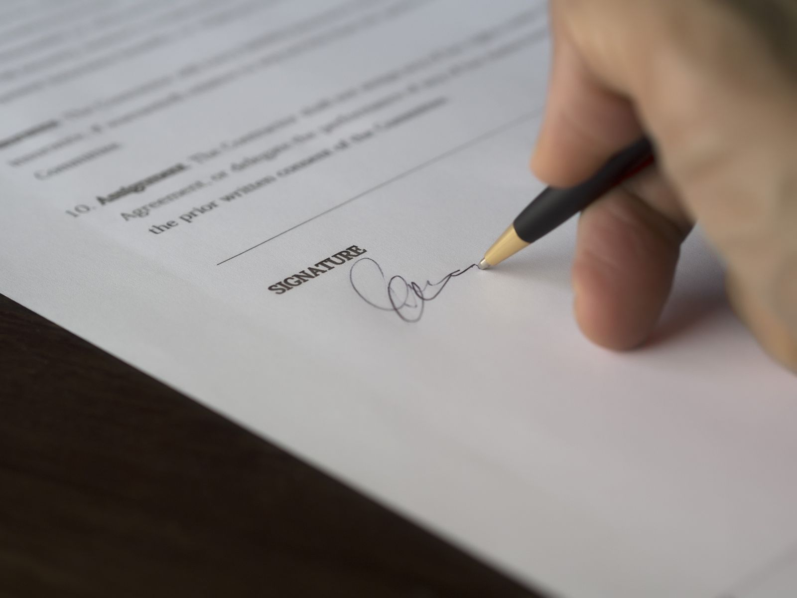 A close-up of a hand signing the signature line of a printed agreement, with an assignment clause visible above it